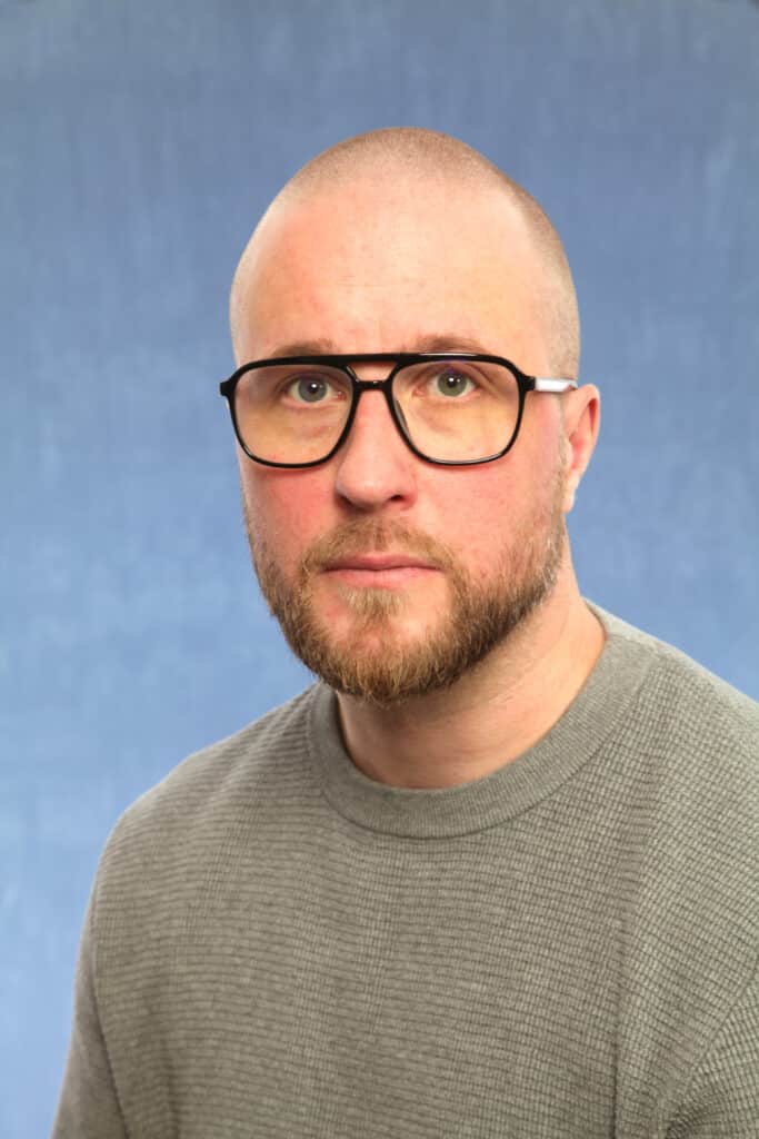Close-up of a bald man with a beard and thick black glasses, wearing a gray sweater, against a blue background.