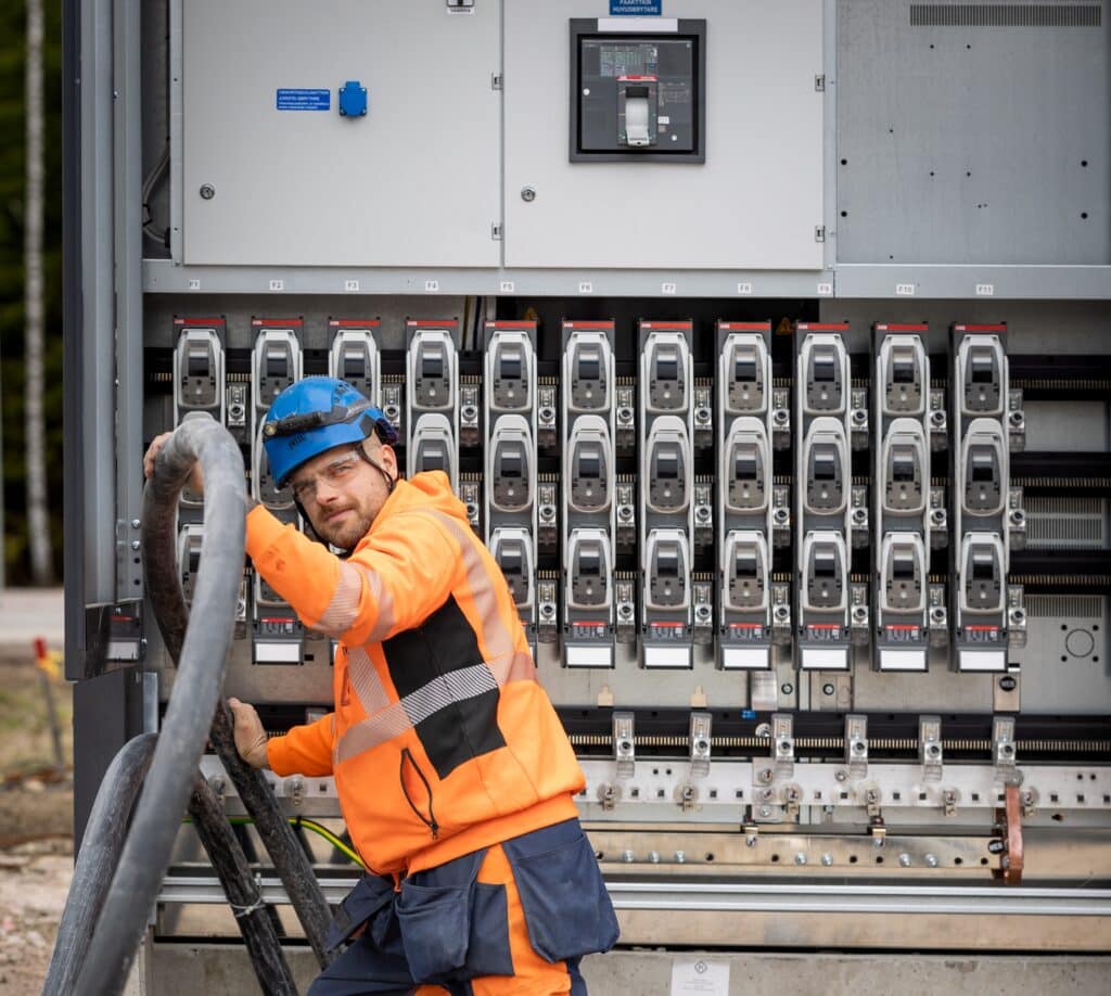 Electrician in an orange safety jacket and blue helmet works on a large outdoor electrical panel with many circuit breakers.