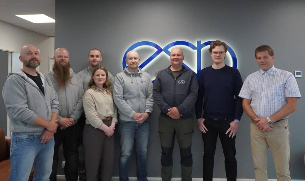 Eight people stand in a line in an office, gray wall behind them with a blue company logo partially visible. (group portrait)