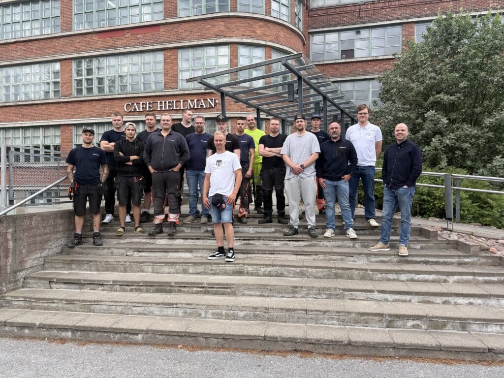 Group of about 20 workers posing on concrete steps in front of a brick building with 'CAFE HELLMAN' sign behind them.