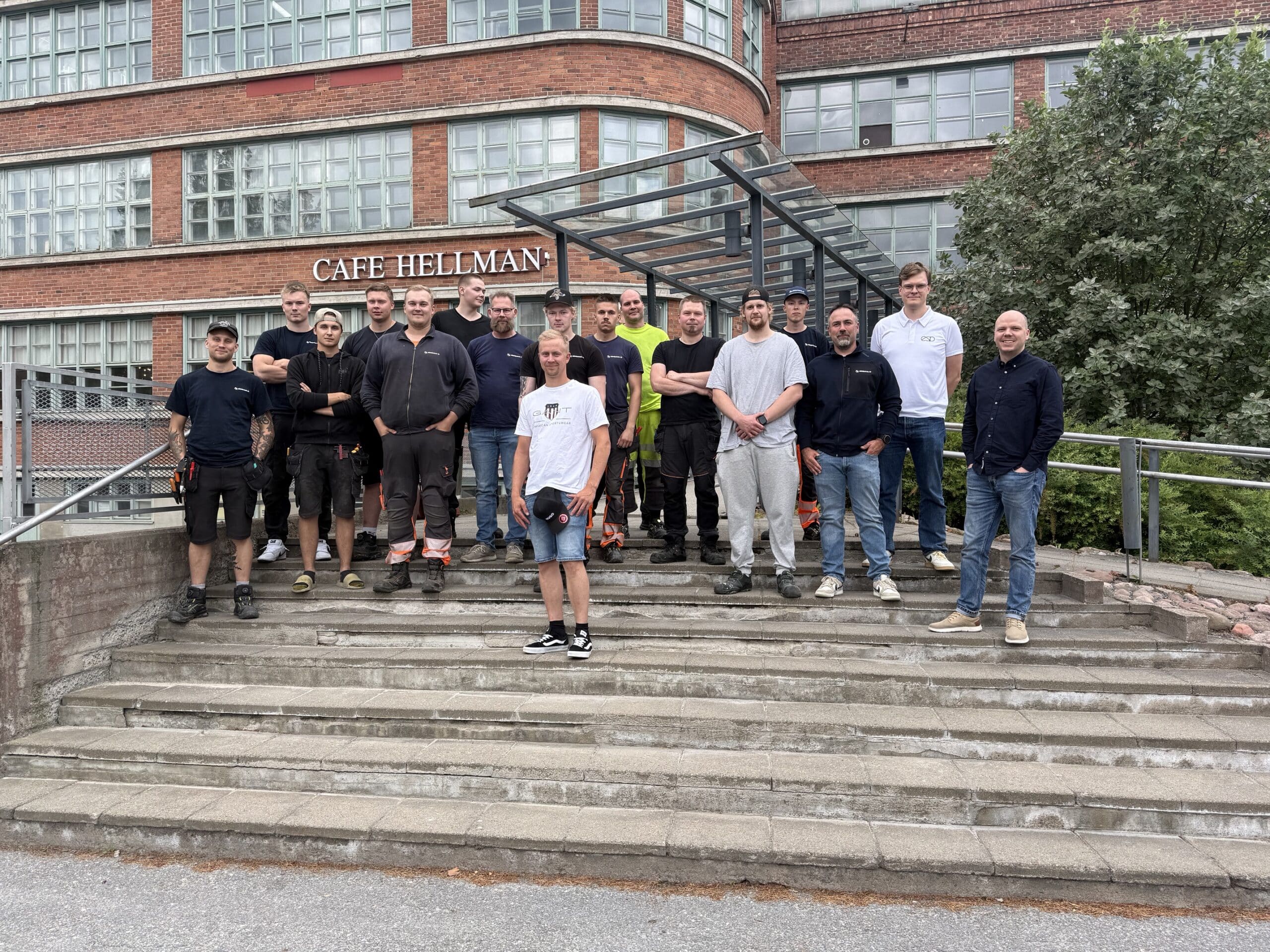 Group of about 20 workers posing on concrete steps in front of a brick building with 'CAFE HELLMAN' sign behind them.